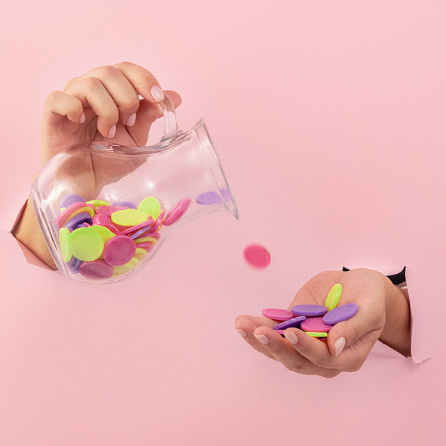 Foto de mãos ultrapassando um parede rosa clara, em que uma delas que segura moedas coloridas e, a outra, uma jarra transparente que despeja os objetos.