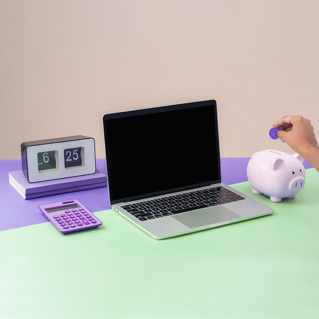 Imagem de um notebook ao lado de uma calculadora roxa, de um cofre de porquinho rosa, e de um livro de capa roxa com um relógio digital sobre ele. Há também uma mão colocando uma moeda dentro do porquinho.