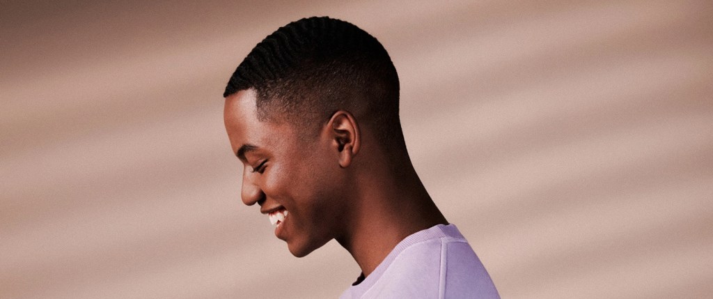 Livelo: Nubank tem parceria com o programa de pontos? Foto mostra um menino negro, de perfil, sorrindo e olhando para baixo. Ele veste uma camiseta de manga longa em tom violeta.