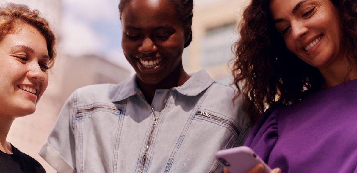 três mulheres sorrindo, uma segurando o celular com uma capinha roxa.