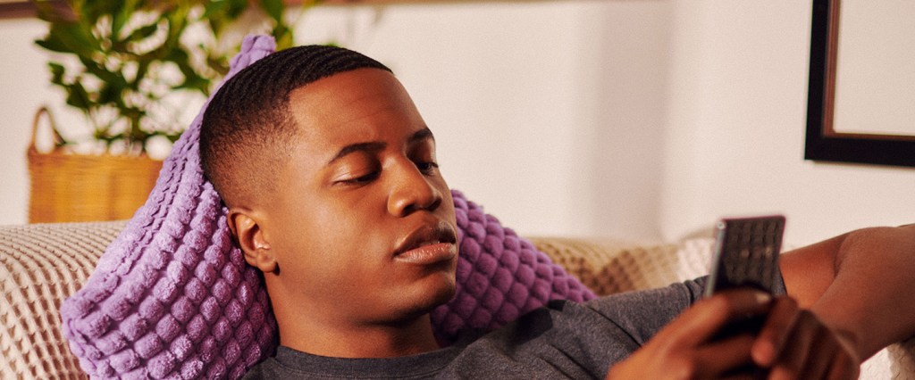 Foto de um homem negro, deitado no sofá com o celular em mãos.
