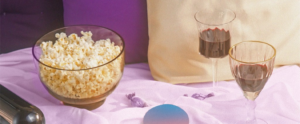 Foto mostra um balde de pipoca e duas taças de vinho tinto em cima de uma cama com lençóis roxos.