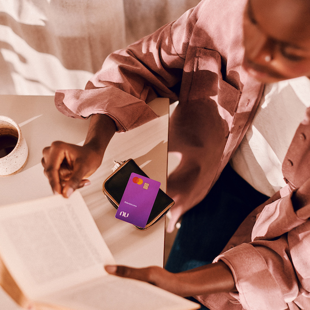 Imagem de uma mulher negra sentada em um café, lendo um livro. Sobre a mesa, está uma carteira com um cartão Nubank. Há também uma xícara de café.