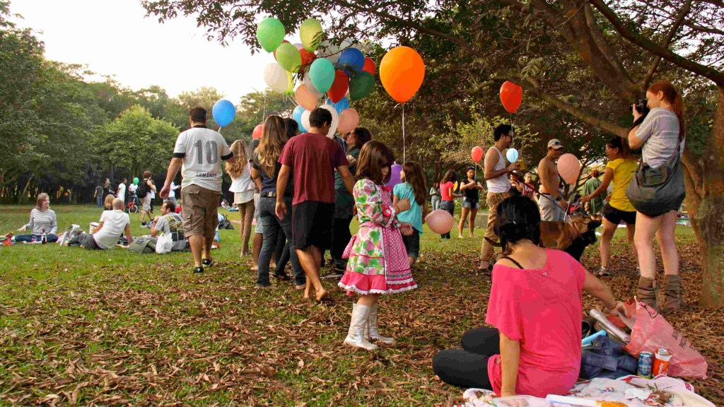 Na imagem aparecem várias pessoas em um parque sob uma árvore segurando balões. Ao centro uma menina de vestido segura um balão laranja e, à direita, uma mulher de rosa está sentada sobre uma toalhe de picnic