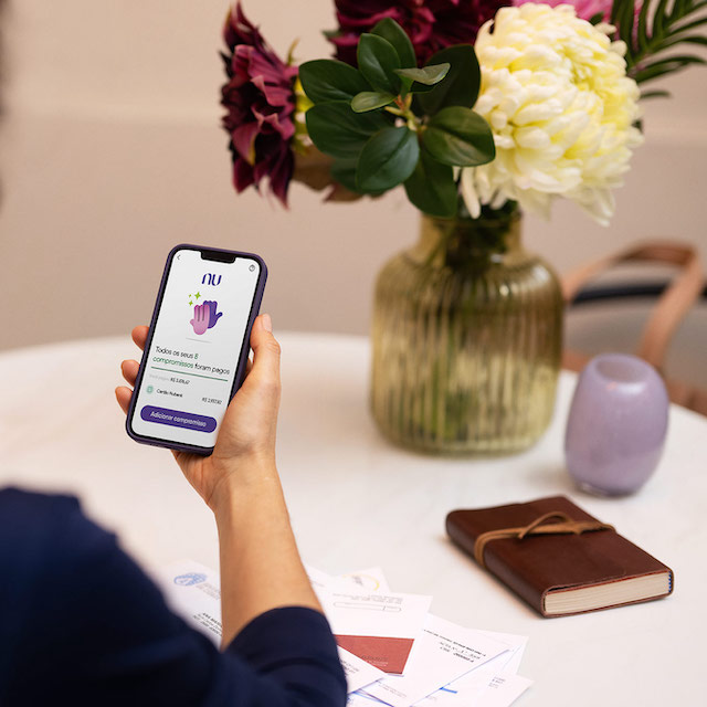 Foto de uma mulher de costas, segurando um celular. Na tela, o app do Nubank com os compromissos financeiros pagos. Um vaso de flor aparece na imagem também.