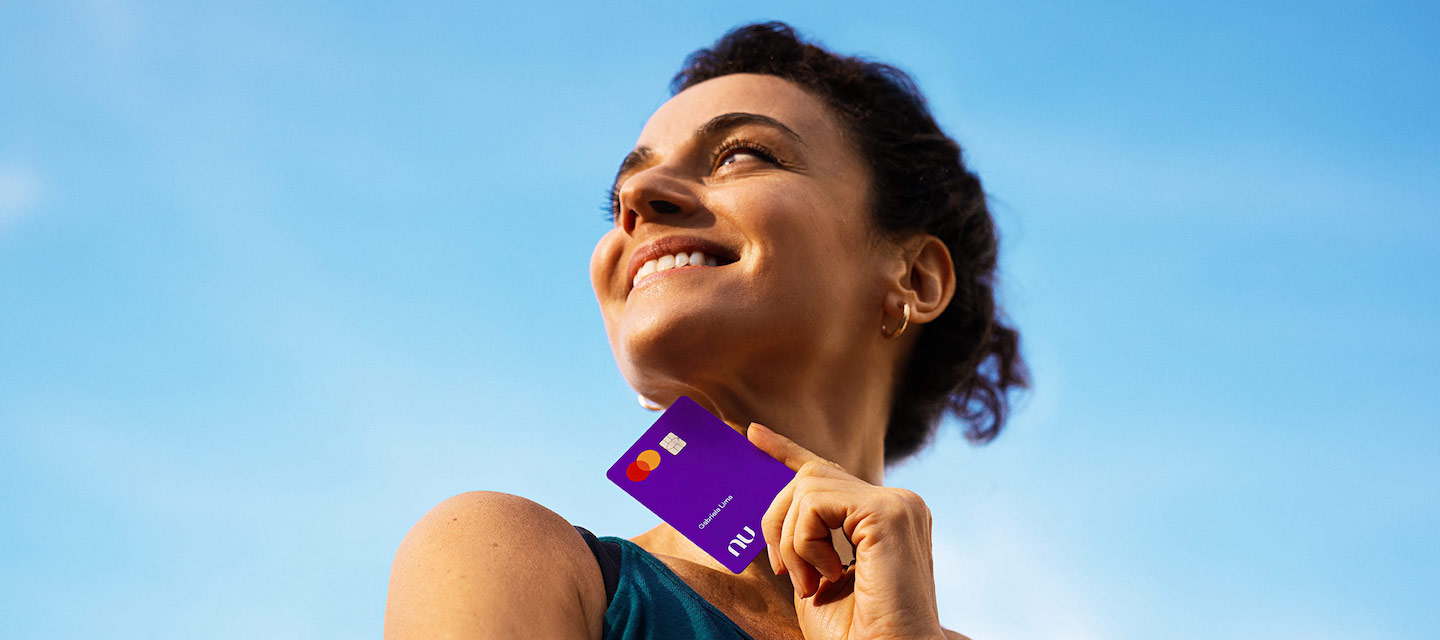 uma mulher branca segurando o cartão Nubank. O céu está azul.