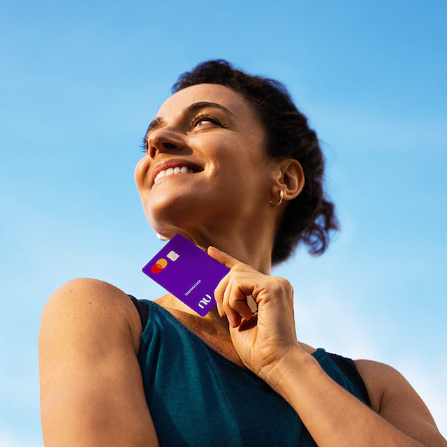 uma mulher branca segurando o cartão Nubank. O céu está azul.