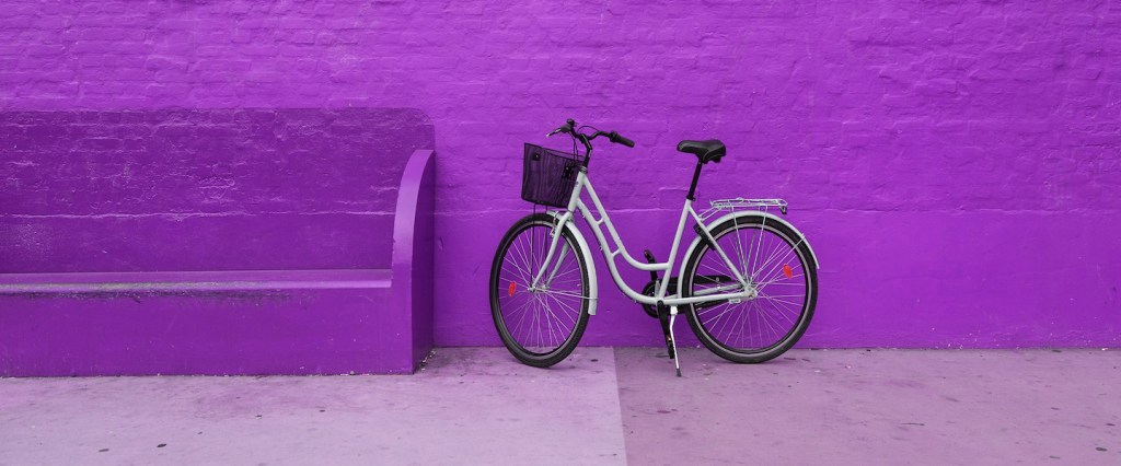 Imagem de uma bicicleta branca, com cestinha preta, encostada em uma parede roxa.