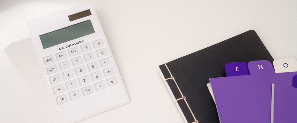 Imagem de uma calculadora branca ao lado de um caderno preto e de etiquetas roxas sobre uma mesa de escritório branca.