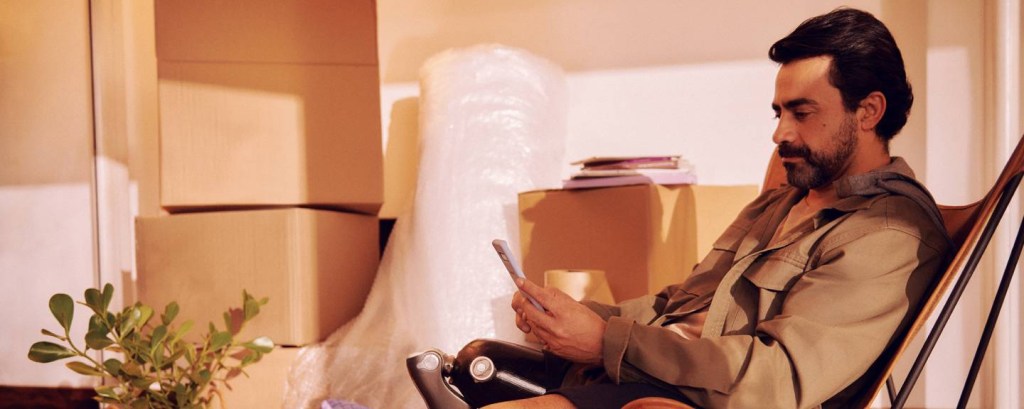 Nubank e B3: imagem mostra um homem de barba e cabelos pretos sentado em uma cadeira em meio a uma sala com várias caixas de mudança. Ele tem uma prótese mecânica na perna direita e está usando o celular. É