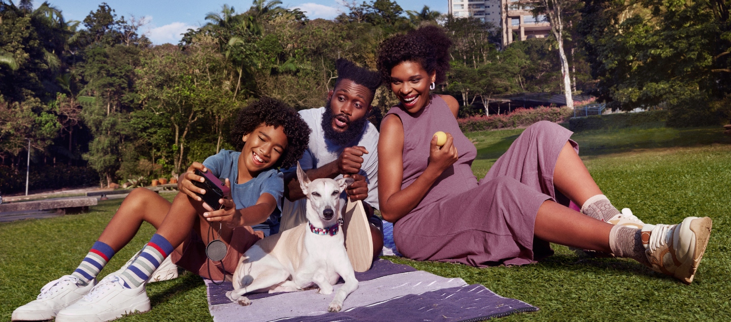 Nubank+ - Imagem de uma família negra, com um cachorro. Eles estão em um parque, sentados numa toalha em tons de roxo.