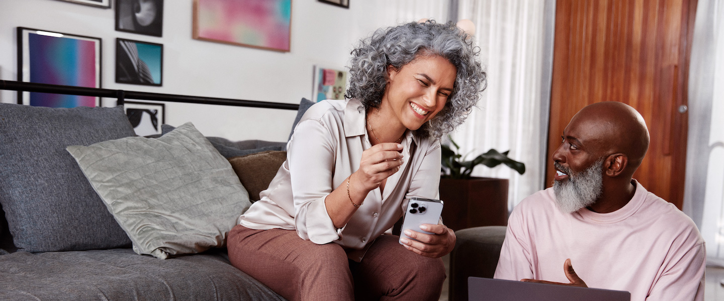 Foto de uma mulher de cabelos brancos encaracolados e um homem negro. Os dois sorrindo e ela mostrando o celular para ele. Ela está sentada no sofá e ele no chão, com um notebook aberto numa mesinha central.