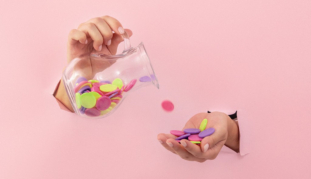 imagem de duas mãos, uma segurando uma jarra derramando moedas coloridas na outra mão. O fundo é rosa claro.