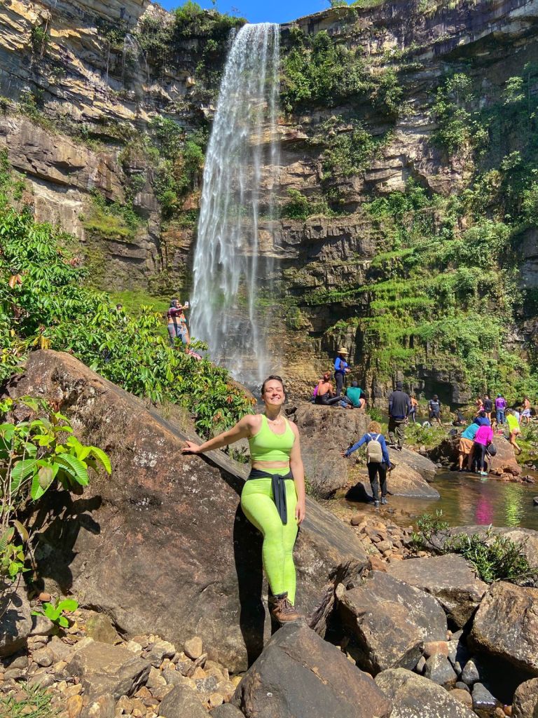 Na foto, Bruna em pé, com um conjunto esporte verde claro, com uma blusa preta amarrada na cintura e uma bota. Ela está encostada num pedra, próxima a uma cachoeira.