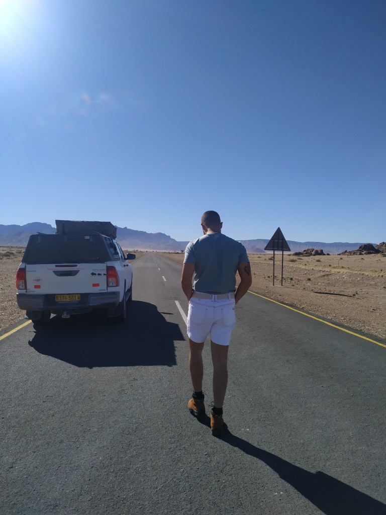 Foto do arquivo pessoa de Pedro, no Parque Nacional Etosha, na Namíbia. Ele no meio da estrada, com uma bermuda jeans e camiseta azul, e seu carro branco em frente.