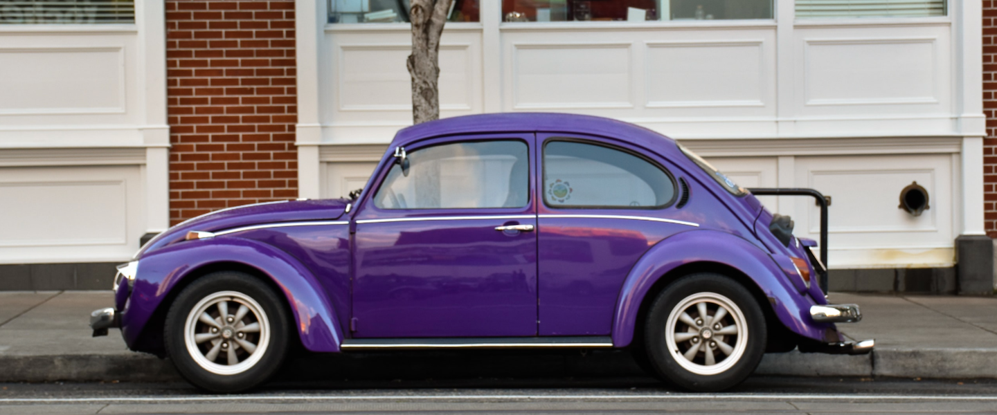 Viagem de carro: como se planejar e evitar surpresas no orçamento. Na imagem, um fusca roxo clássico, conservado e pouco modificado em uma rua estacionado na vaga. Ao fundo, um imóvel com janelas brancas grandes fechadas e tijolos marrons entre elas, além do tronco de uma árvore.