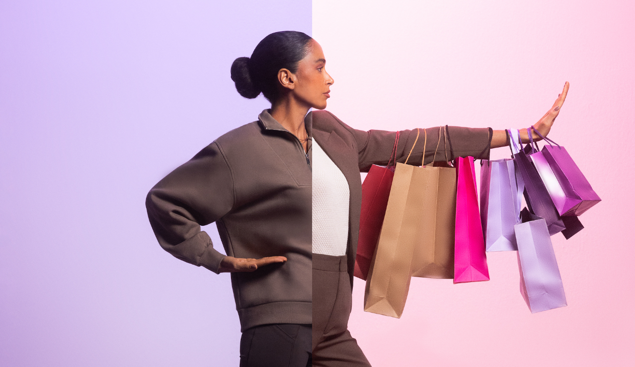 Na imagem, uma mulher negra, com cabelo preso em coque, veste roupas em marrom e está posicionada ao centro, com o braço direito esticado em direção ao lado direito na foto. Nele, a mulher carrega diversas sacolas de compras em tons de roxo, rosa e bege.