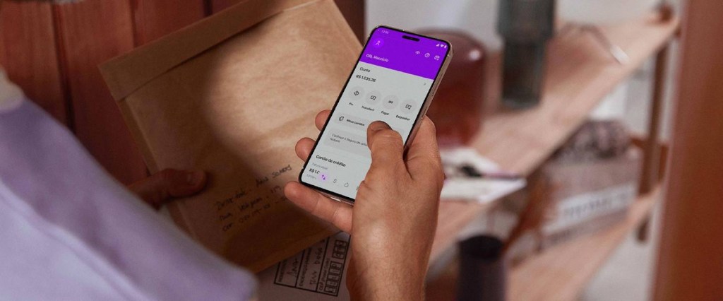 Compra programada de criptomoedas - foto mostra a mão de uma pessoa usando um celular. Na tela, é possível ver o app do Nubank.