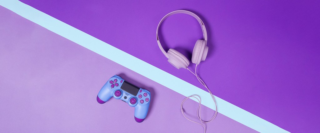 Imagem mostra um controle de videogame e um fone de ouvido, ambos em tons de roxo e rosa, em cima de uma mesa, também roxa com uma faixa azul na diagonal.