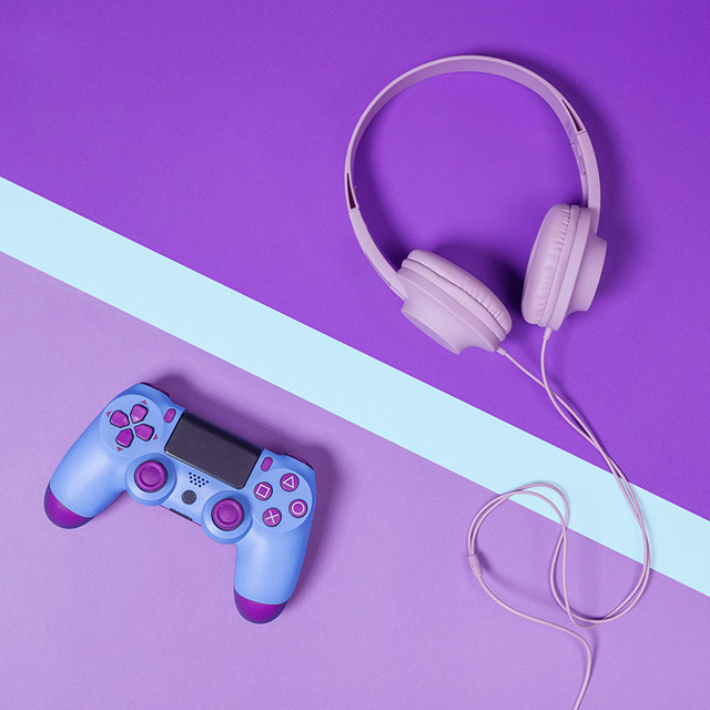 Imagem mostra um controle de videogame e um fone de ouvido, ambos em tons de roxo e rosa, em cima de uma mesa, também roxa com uma faixa azul na diagonal.