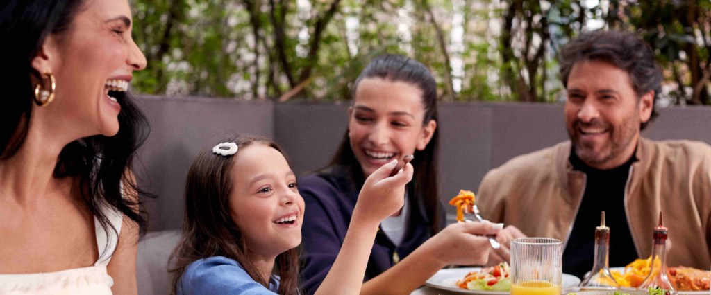 Espaço Família no app Nubank: na imagem, uma família está sentada à mesa, fazendo uma refeição. Ao lado esquerdo da foto, uma mulher branca de cabelos escuros e lisos sorri. Ao centro, uma criança branca de cabelos castanhos e uma adolescente branca de cabelos castanhos se olham e sorriem. No canto direito, um homem branco de cabelos castanhos, barba e bigode olha para elas.