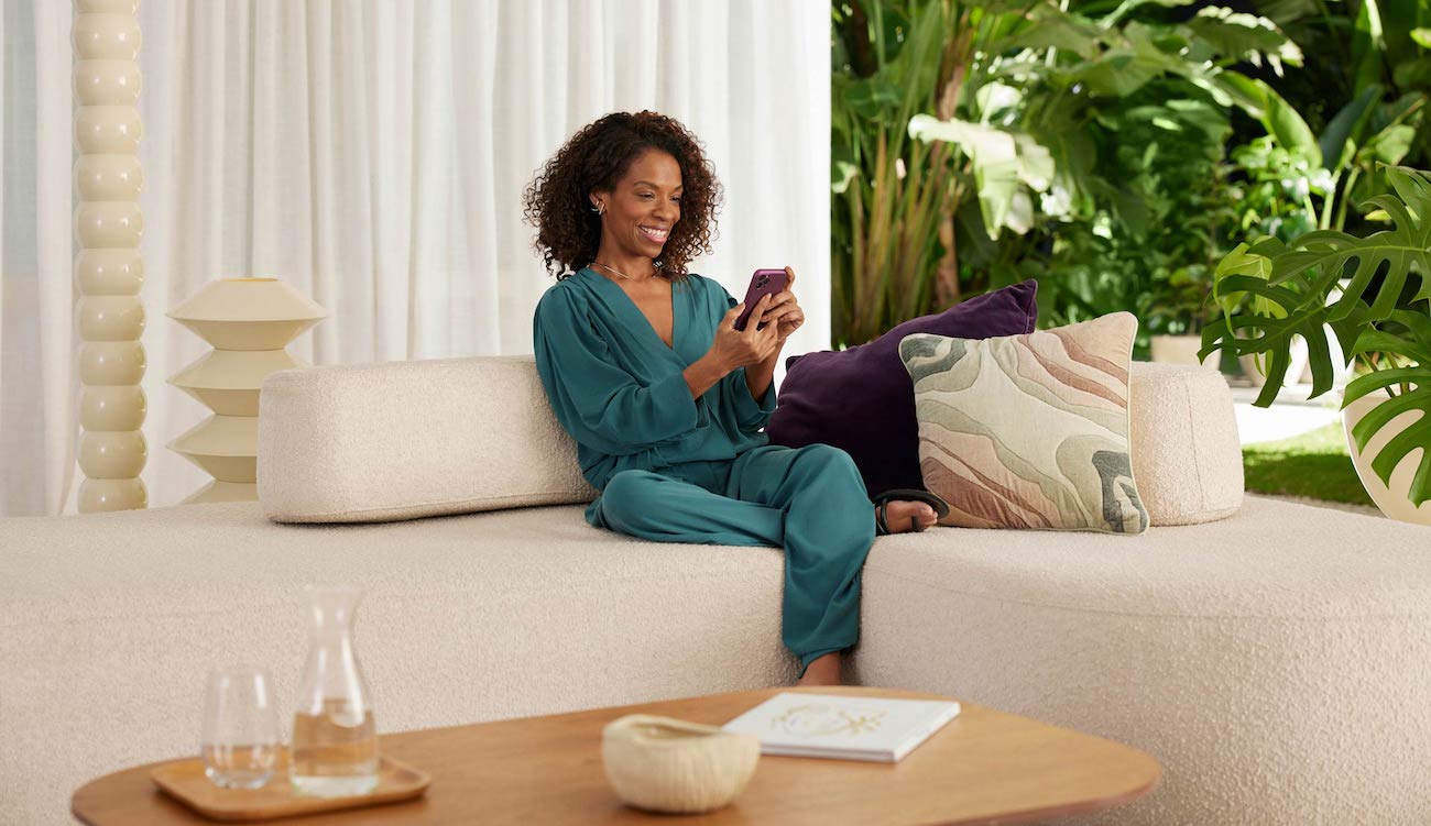 Nubank inicia testes com inteligência artificial generativa: imagem de uma mulher usando uma roupa verde escura. Ela está sentada em um sofa claro, em uma sala com plantas atrás. Está usando um celular de capa roxa.