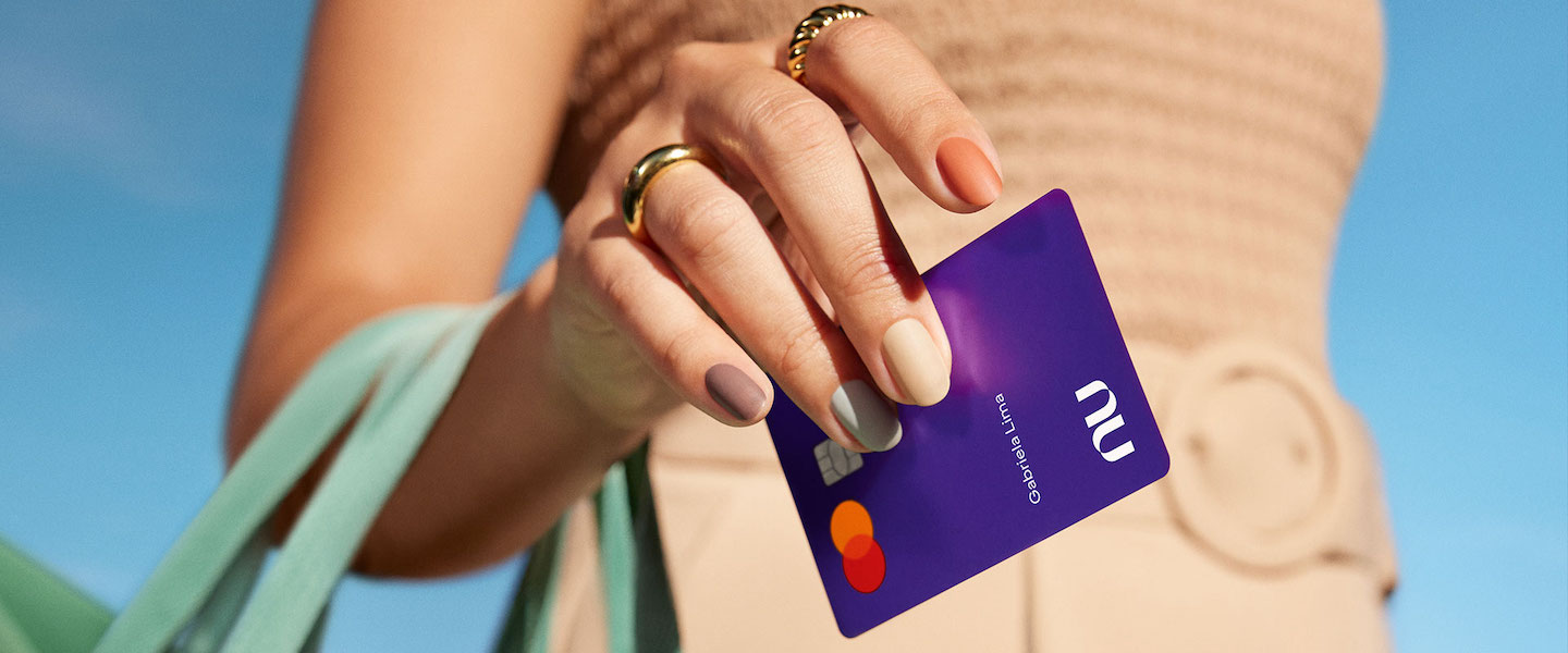 Nubank recebe classificação da Moody's. Foto com close na mão de uma mulher segurando uma bolsa e um cartão Nubank. Ela tem anéis e usa roupas bege.