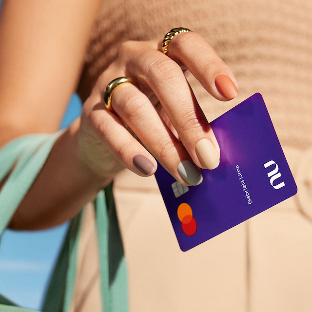 Nubank recebe classificação da Moody's. Foto com close na mão de uma mulher segurando uma bolsa e um cartão Nubank. Ela tem anéis e usa roupas bege.