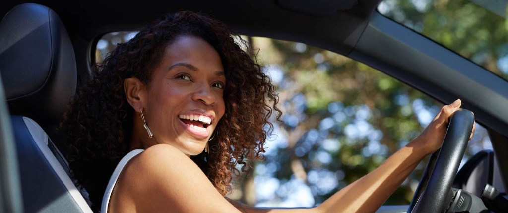 Foto mostra uma mulher negra, de cabelos cacheados acobreados, sorrindo ao volante de um carro. Ela está de regata branca e e um grande brinco prata.