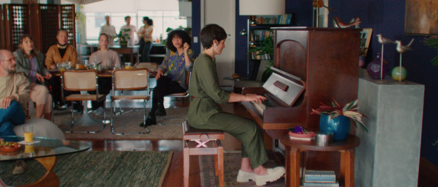 Nubank e Spotify - imagem mostra uma mulher branca de cabelos curtos tocando um piano de madeira em uma grande sala. Ela veste um macacão verde e sapatos brancos. Outras pessoas a observam tocar.