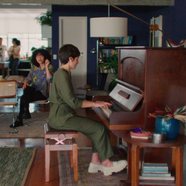 Nubank e Spotify - imagem mostra uma mulher branca de cabelos curtos tocando um piano de madeira em uma grande sala. Ela veste um macacão verde e sapatos brancos. Outras pessoas a observam tocar.