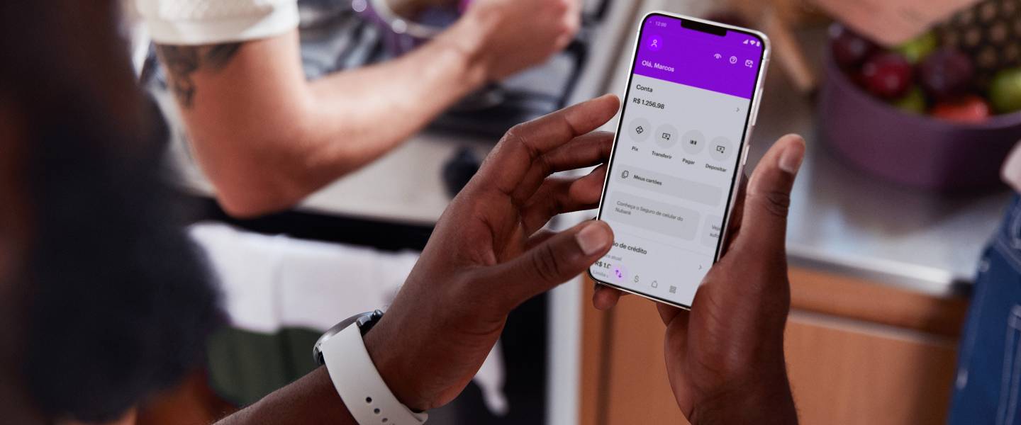 Imagem mostra as mãos de uma pessoa negra segurando um celular. Ela também usa um relógio branco. Na tela do celular está possível ver o app do Nubank. Ao fundo, duas pessoas estão em uma cozinha, e uma delas mexe uma panela roxa que está no fogão.