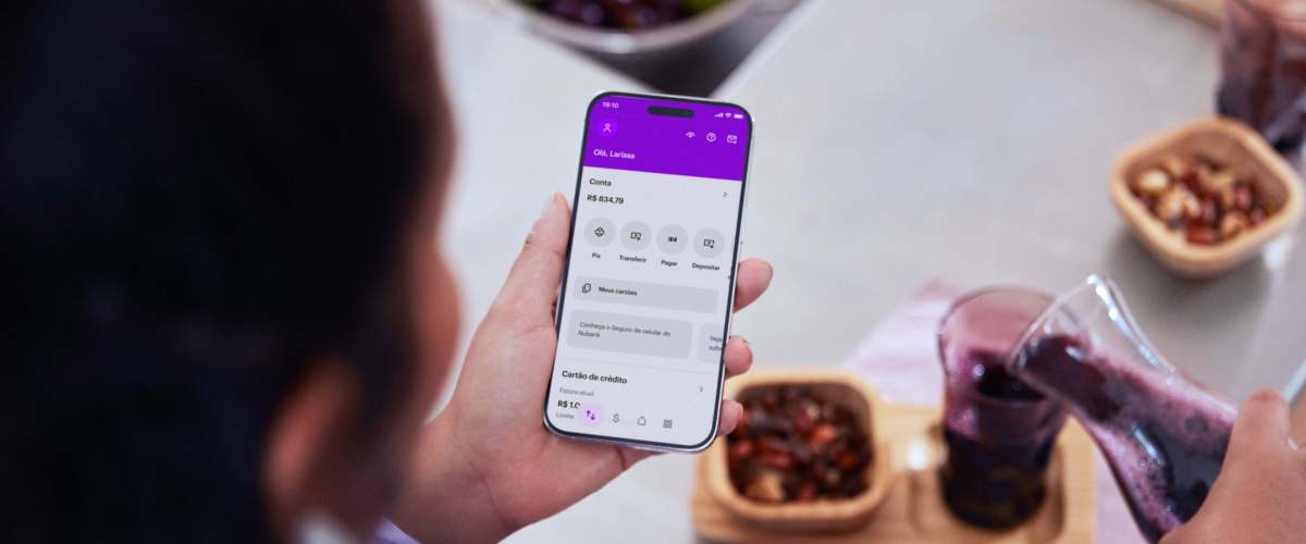 Na imagem aparece a mão de uma muler branca segurando um celular com o aplicativo do Nu aberto. Ao fundo, uma mesa de café da manhã com frutas e suco roxo.