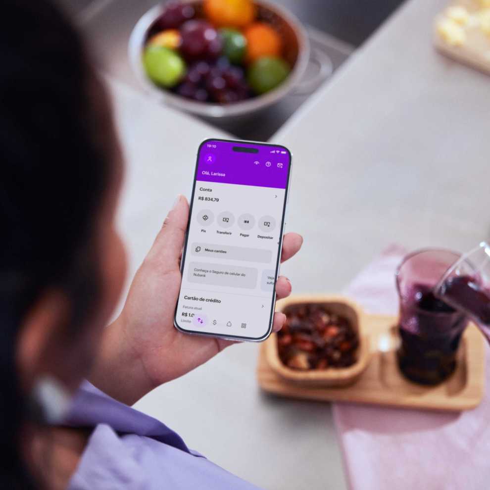 Na imagem aparece a mão de uma muler branca segurando um celular com o aplicativo do Nu aberto. Ao fundo, uma mesa de café da manhã com frutas e suco roxo.