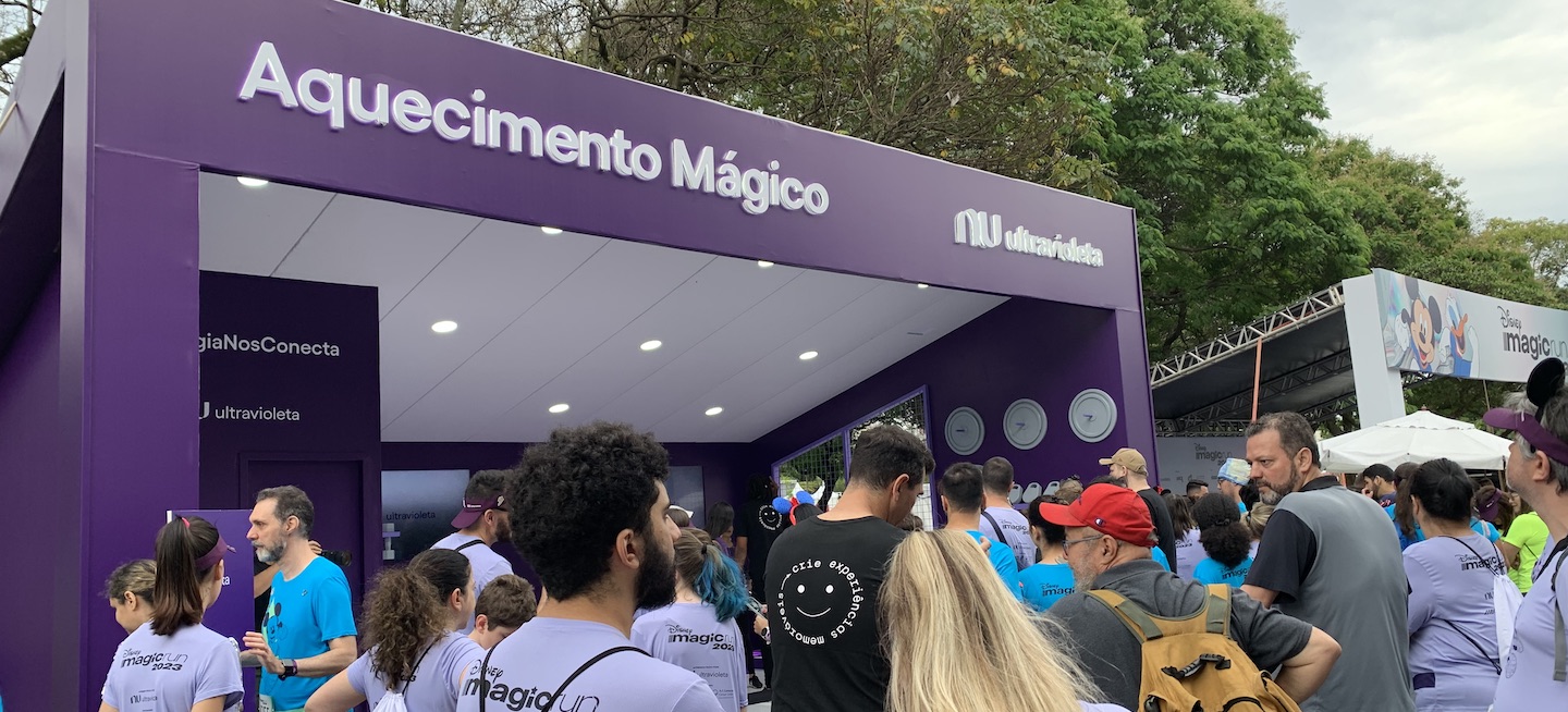 Foto mostra diversas pessoas numa fila em frente ao estande aquecimento mágico do Nubank na Disney Magic Run. Elas vestem a camiseta da corrida, roxa clara. O estande é roxo escuro.