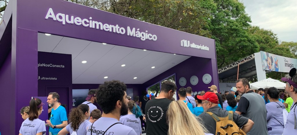 Foto mostra diversas pessoas numa fila em frente ao estande aquecimento mágico do Nubank na Disney Magic Run. Elas vestem a camiseta da corrida, roxa clara. O estande é roxo escuro.