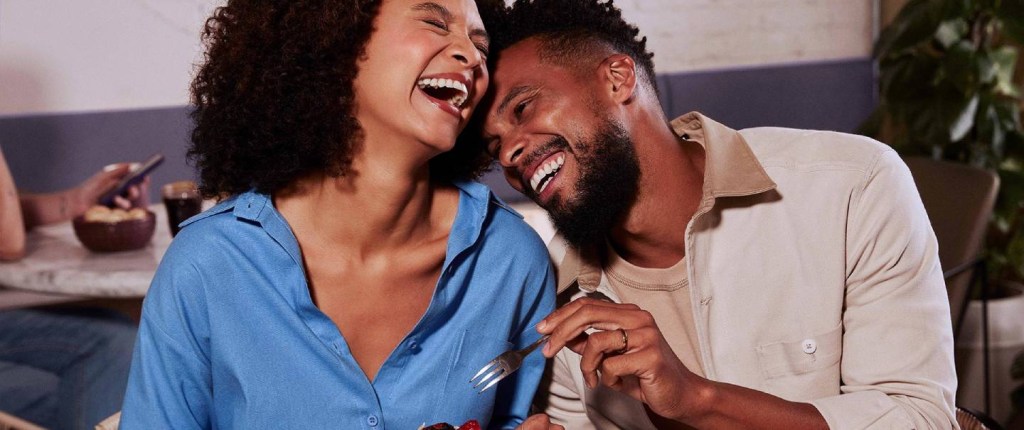 Nucoin - imagem mostra uma mulher e um homem negros sorrindo. A mulher veste uma camisa azul e o homem uma camiseta e um casaco beges. Eles estão próximos um do outro e sorriem. A mulher tem um bolo de chocolate nas mãos e o homem segura um garfo.