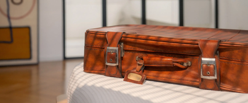 imagem de uma mala de viagem sobre uma cama com lençol branco. A mala é marrom, de couro.