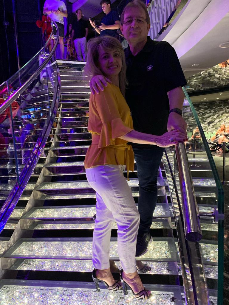 O casal Rosana e João Francisco Favoreto em uma escada brilhante de um navio e cruzeiro. Ela usa uma calça branca e blusa amarela. Ele, uma calça jeans e uma blusa preta.