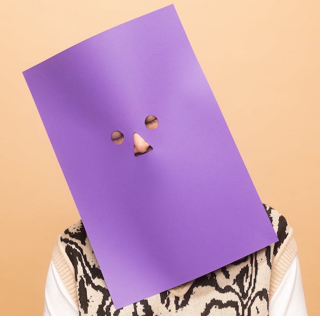 Imagem mostra uma pessoa com umas máscara de papel roxa no rosto. Ela está com os olhos fechados. O fundo da foto é laranja.