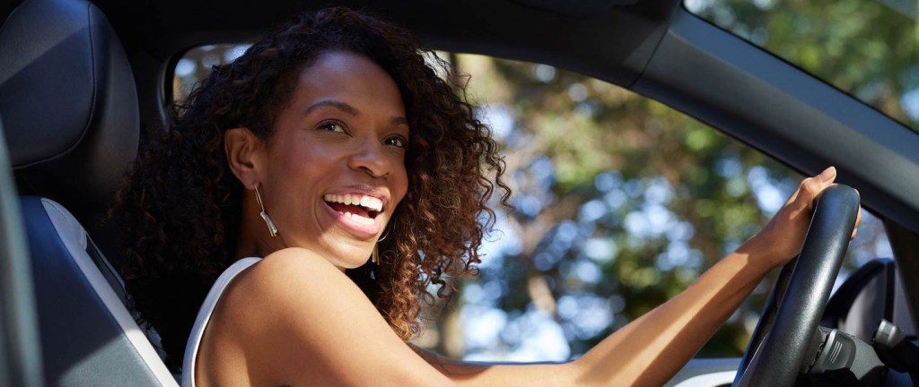 Foto mostra uma mulher negra, de cabelos cacheados acobreados, sorrindo ao volante de um carro. Ela está de regata branca e usa um grande brinco prata.