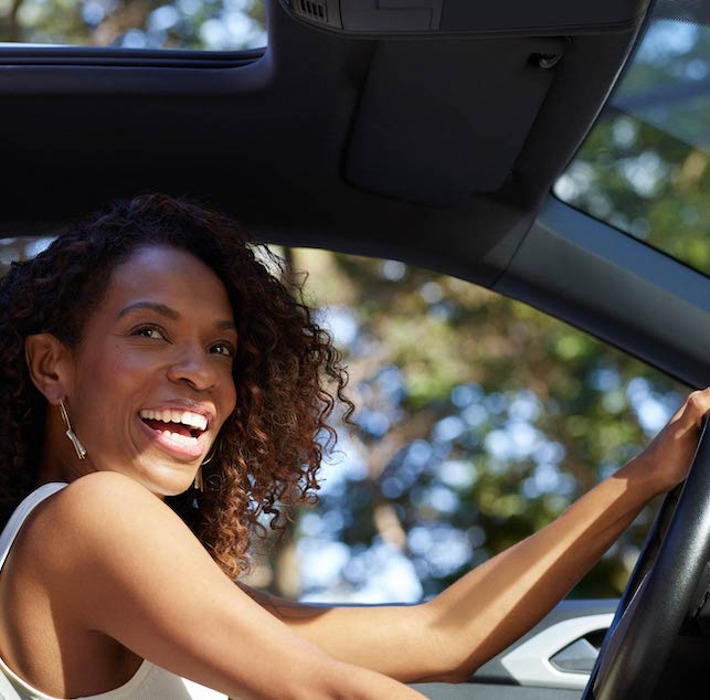 Foto mostra uma mulher negra, de cabelos cacheados acobreados, sorrindo ao volante de um carro. Ela está de regata branca e usa um grande brinco prata.