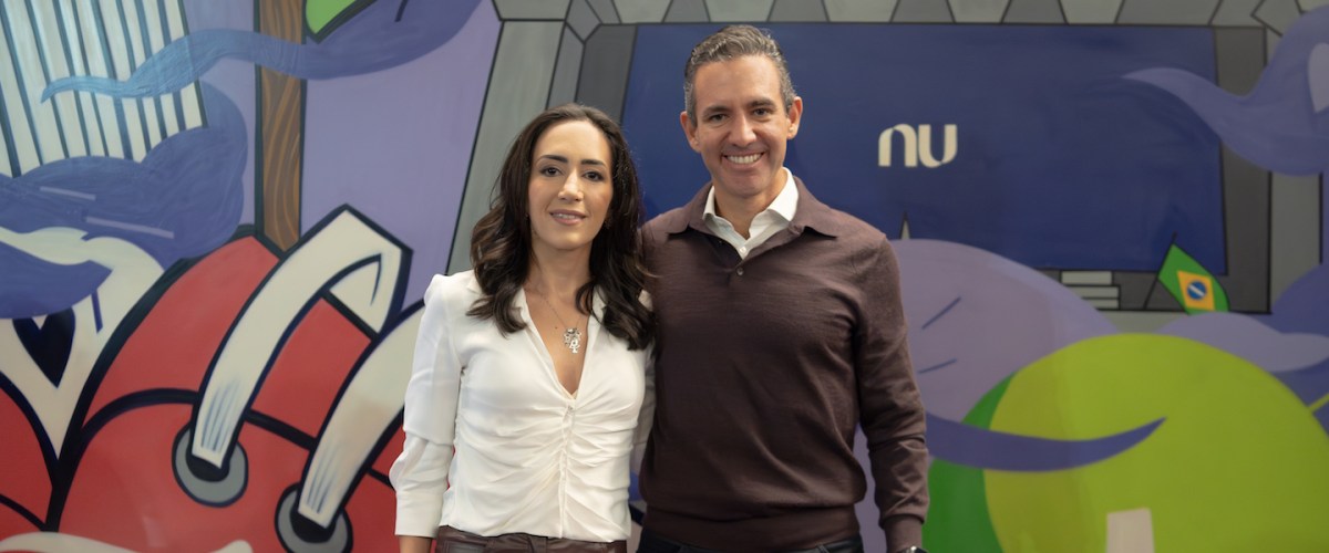 David Velez e Cristina Junqueira posam em frente a uma parede grafitada com o logo do Nubank ao fundo