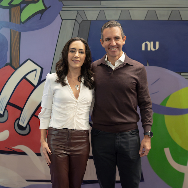 David Velez e Cristina Junqueira posam em frente a uma parede grafitada com o logo do Nubank ao fundo