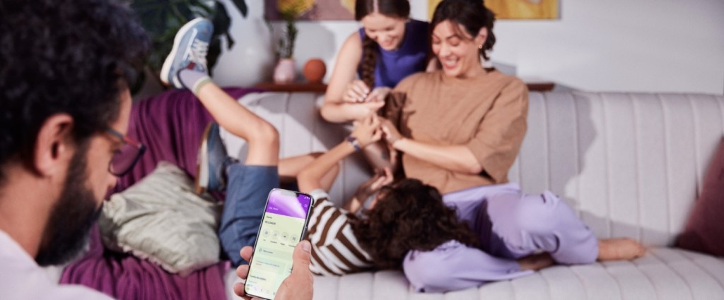 Nubank+ - Quatro pessoas em uma sala, em primeiro plano um homem de costas segura um celular em sua mão direita enquanto visualiza sua conta no aplicativo do Nubank, ao fundo há uma mulher sentada no sofá e um garoto deitado com a cabeça em seu colo, atrás do sofá uma garota sorrindo interage com eles.