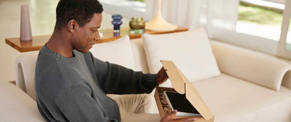 Foto mostra um homem negro, de cabelo escuro e curto, vestido com uma malha cinza, sentado num sofá branco. Ele abre uma caixa de papelão. Dentro dela há um tablet.