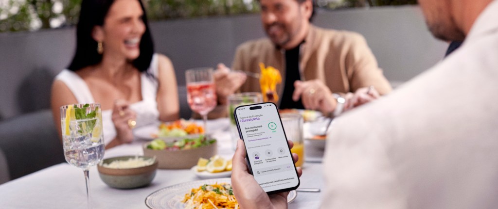 foto mostra três pessoas brancas, com roupas claras, sentadas em um restaurante. No primeiro plano, um homem, de costas, segura um celular. Na tela, o app do Nubank está aberto.