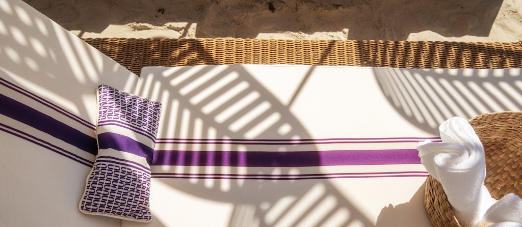 Foto mostra uma esteira na praia com uma toalha branca e detalhes roxos.