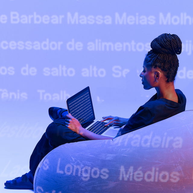 Imagem de uma mulher negra de cabelos trançados presos, sentada em em puff com um notebook em mãos. Ao fundo, tom roxo e várias palavras escritas em branco.