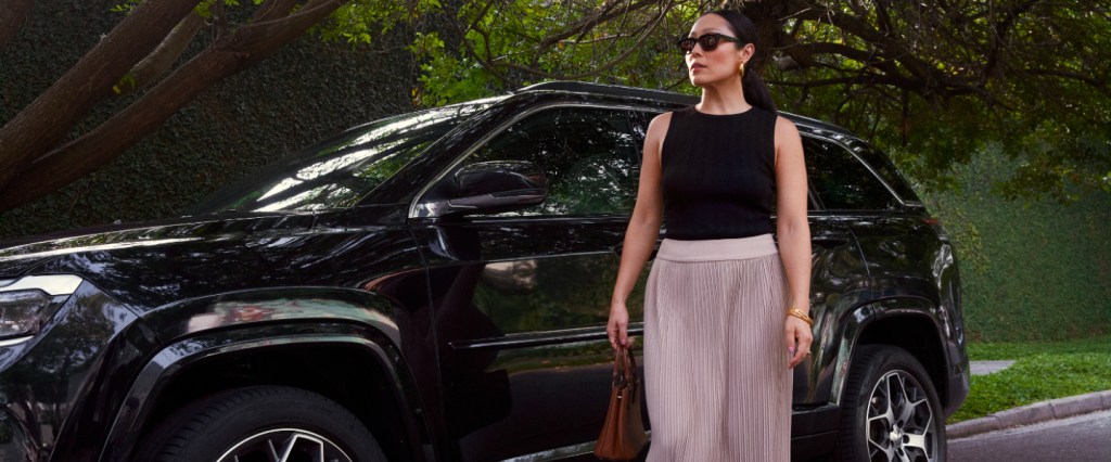 Contas do Detran Nubank: na imagem, uma mulher branca está em pé, em frente a um carro SUV preto, estacionado em uma rua com árvores. Ela está vestindo uma calça rosa claro, uma blusa preta sem mangas, segurando uma bolsa cor marrom na mão esquerda. Ela tem os cabelos lisos e pretos, presos em um rabo de cavalo e usa um óculos de sol preto no rosto.